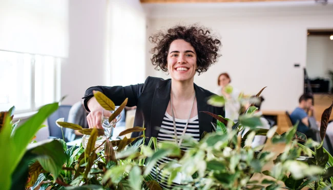Eine Frau gießt Pflanzen im Büro und lächelt in die Kamera.