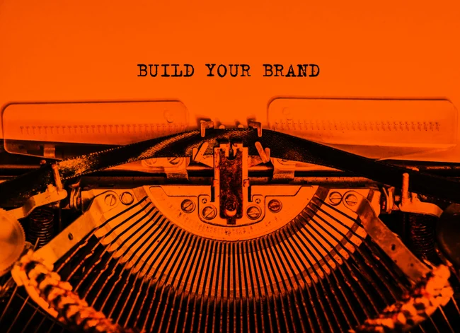 Eine Schreibmaschine in orange auf orangem Hintergrund, die "BUILD YOUR BRAND" auf ein oranges Stück Papier geschrieben hat.