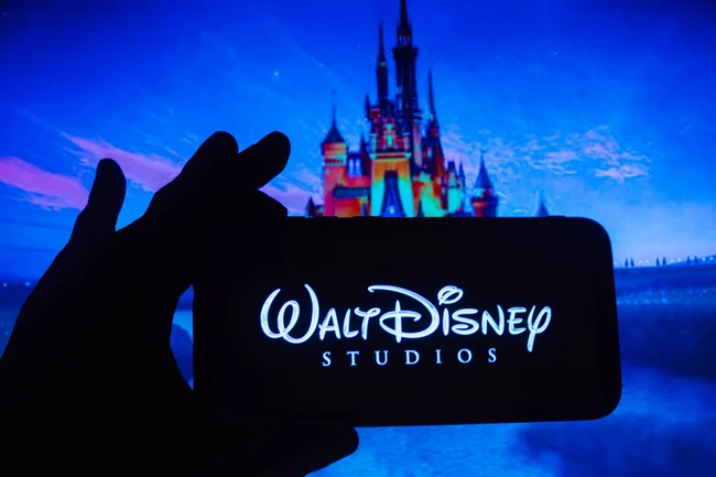 Das Walt Disney Logo auf einem Smartphone wird vor das Disney Schloss gehalten.
